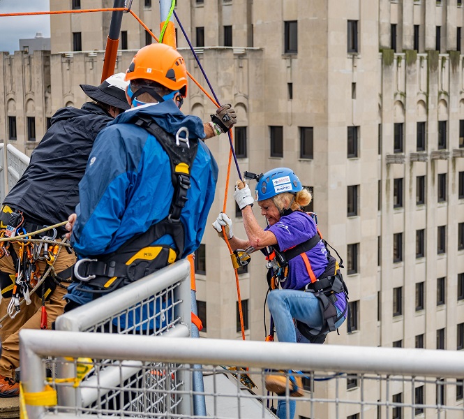 People in helmets and harnesses prepare to rappel down a tall building.