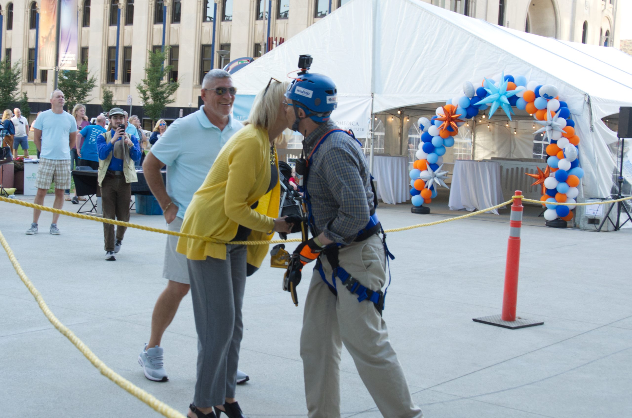 Man in climbing gear and helmet kisses woman in yellow outdoors.