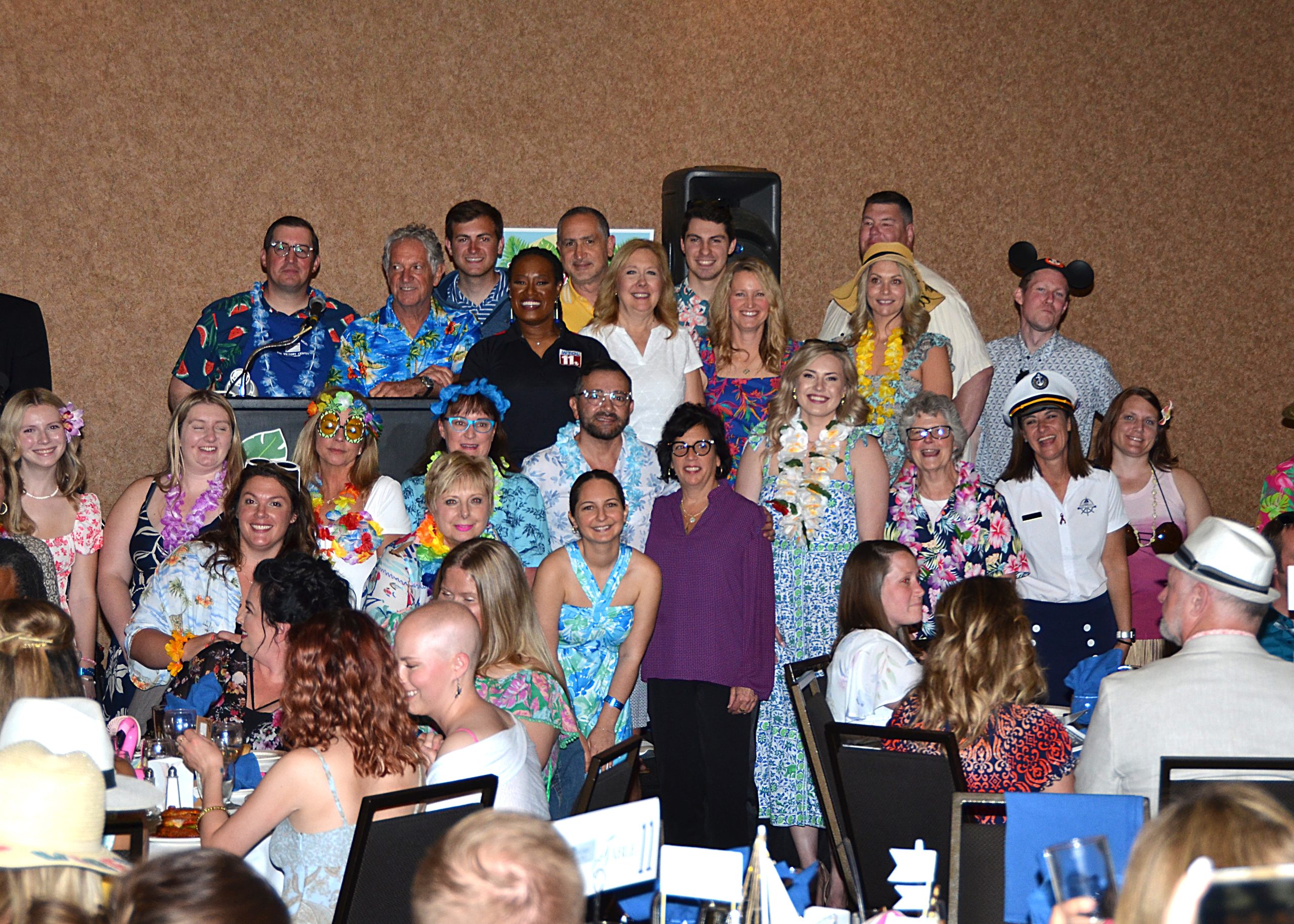 Group of people smiling, wearing colorful, tropical-themed attire in a banquet setting.