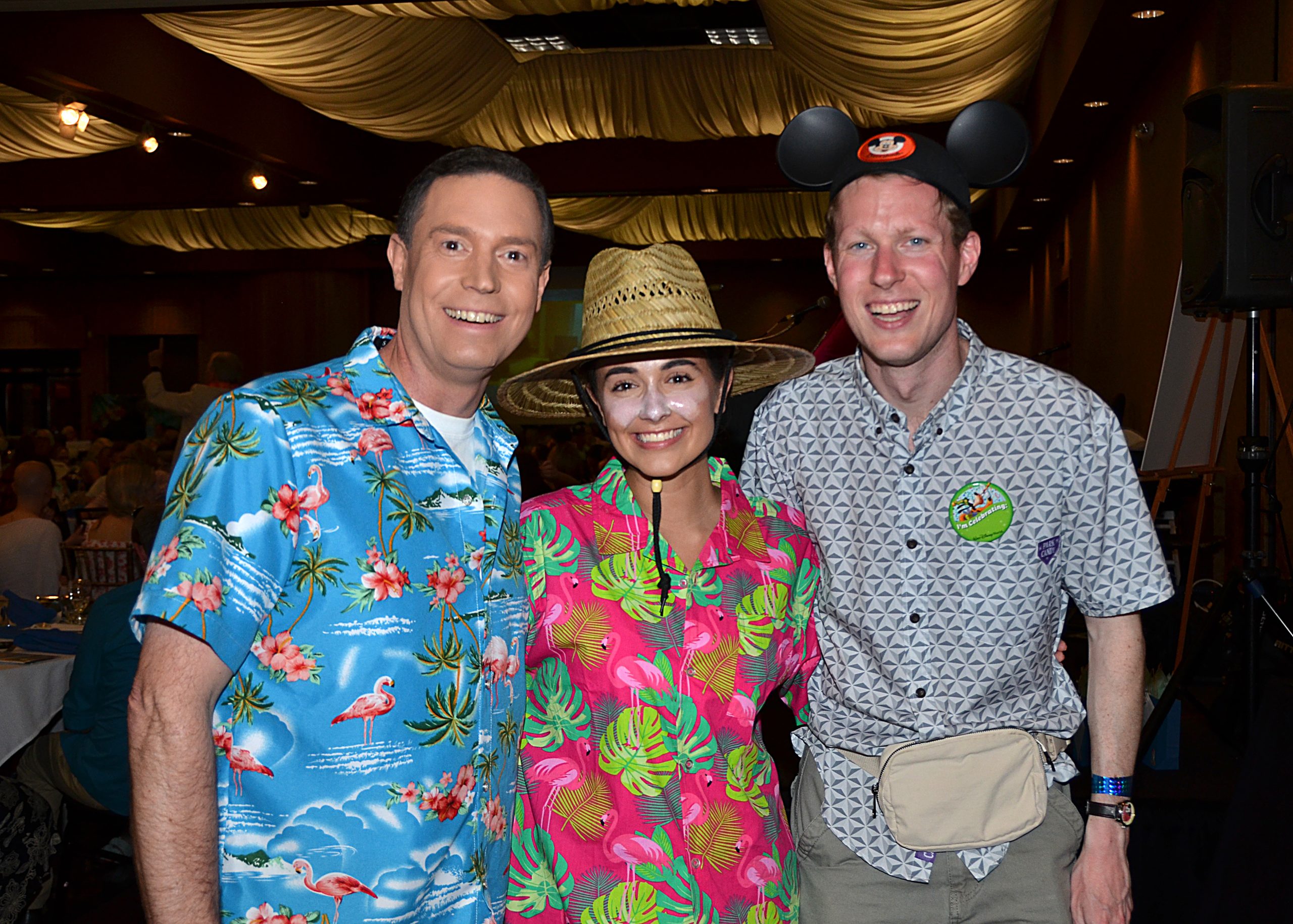 Three people in colorful shirts smiling; one wears a straw hat, another has mouse ears.