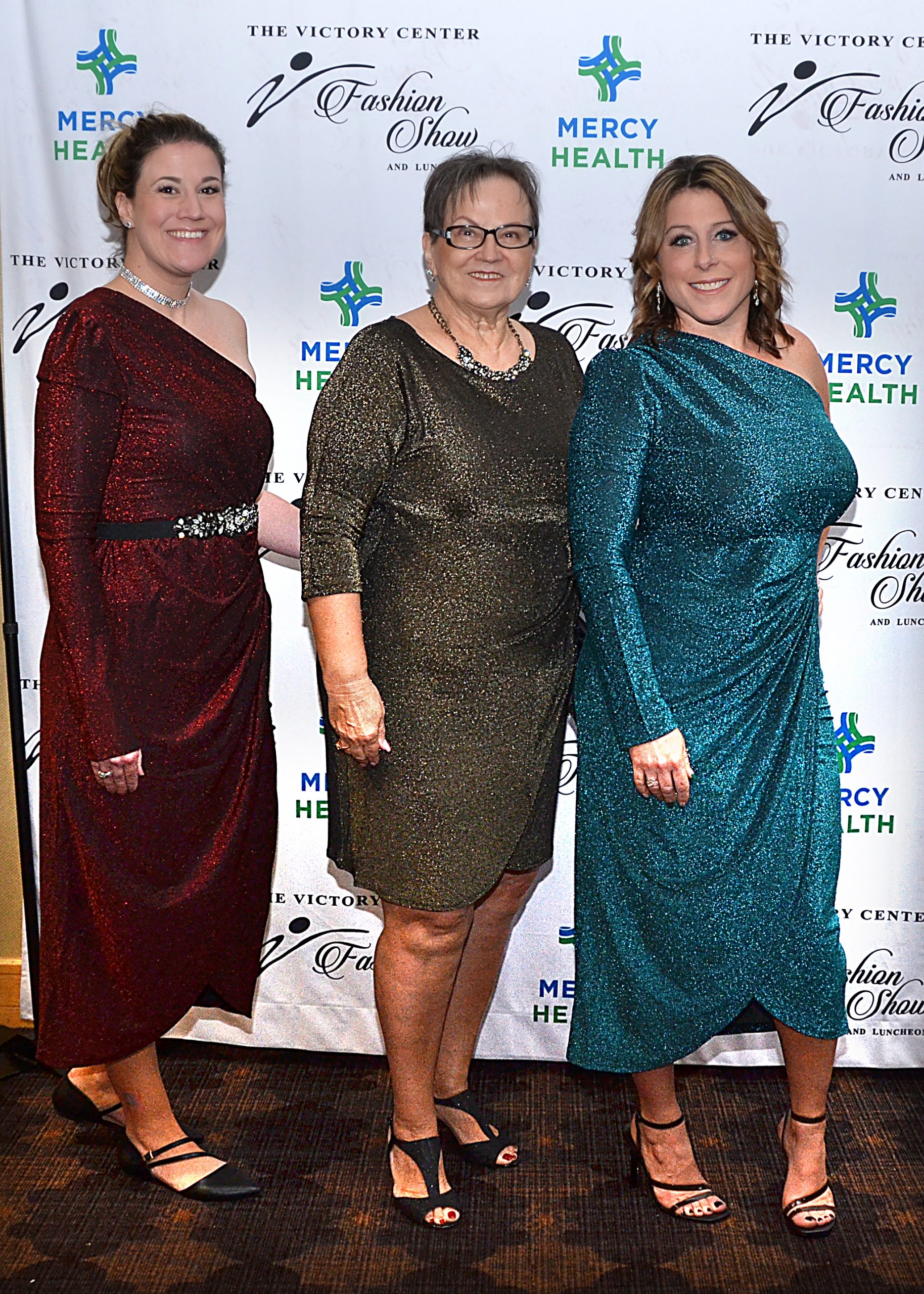 Three women in evening gowns stand smiling in front of a Mercy Health backdrop.