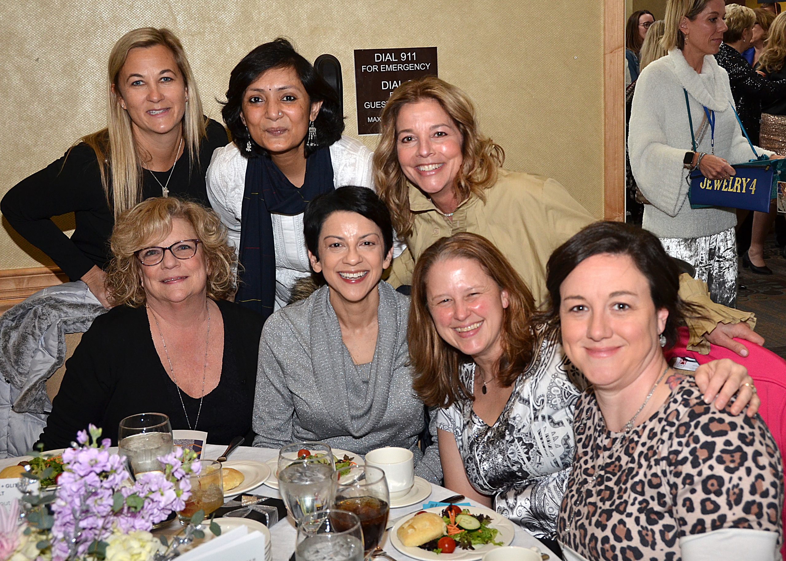 Group of seven women smiling at a dining table with food and drinks.