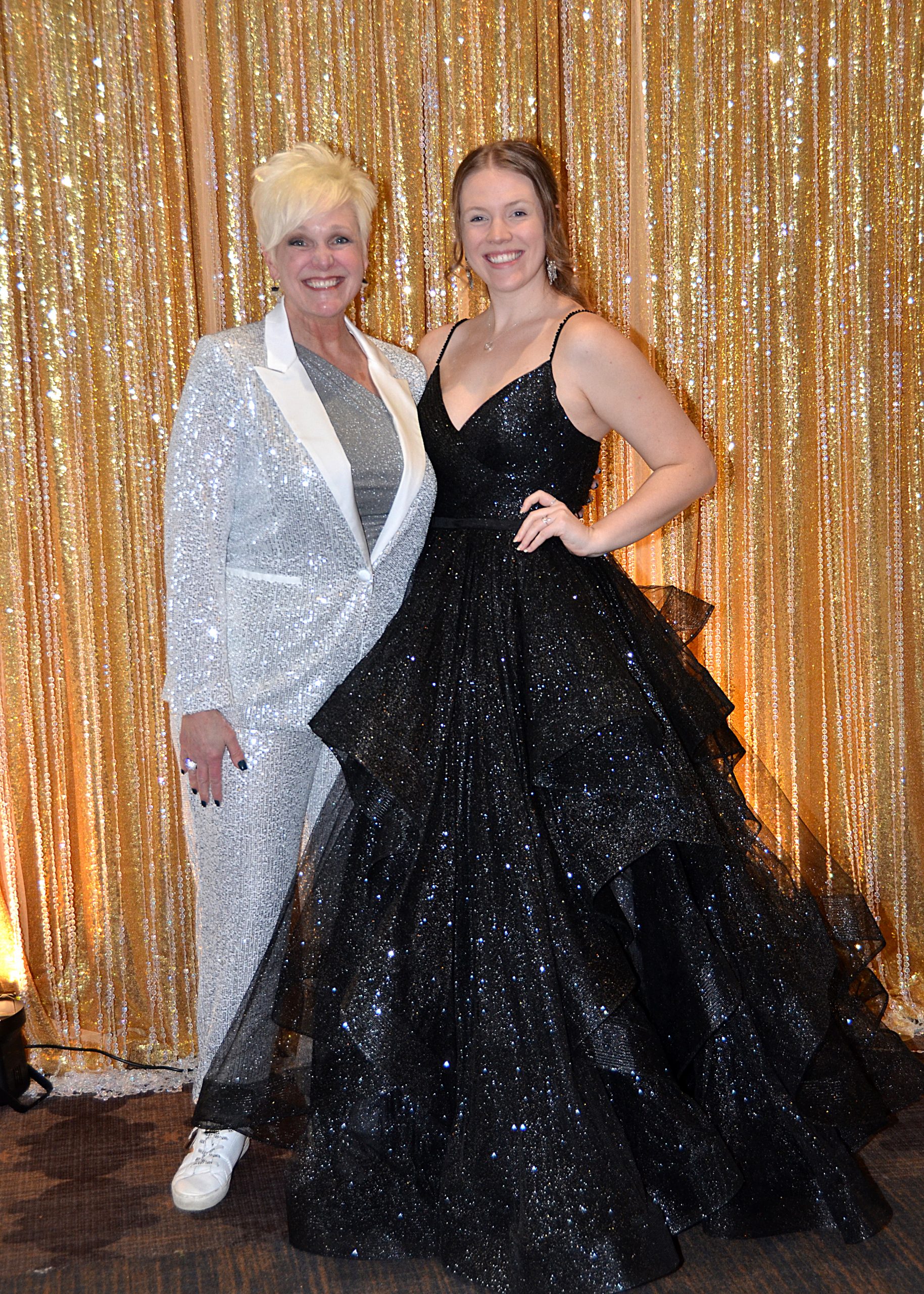 Two women smiling, one in a silver suit, the other in a black gown, standing before a gold curtain.