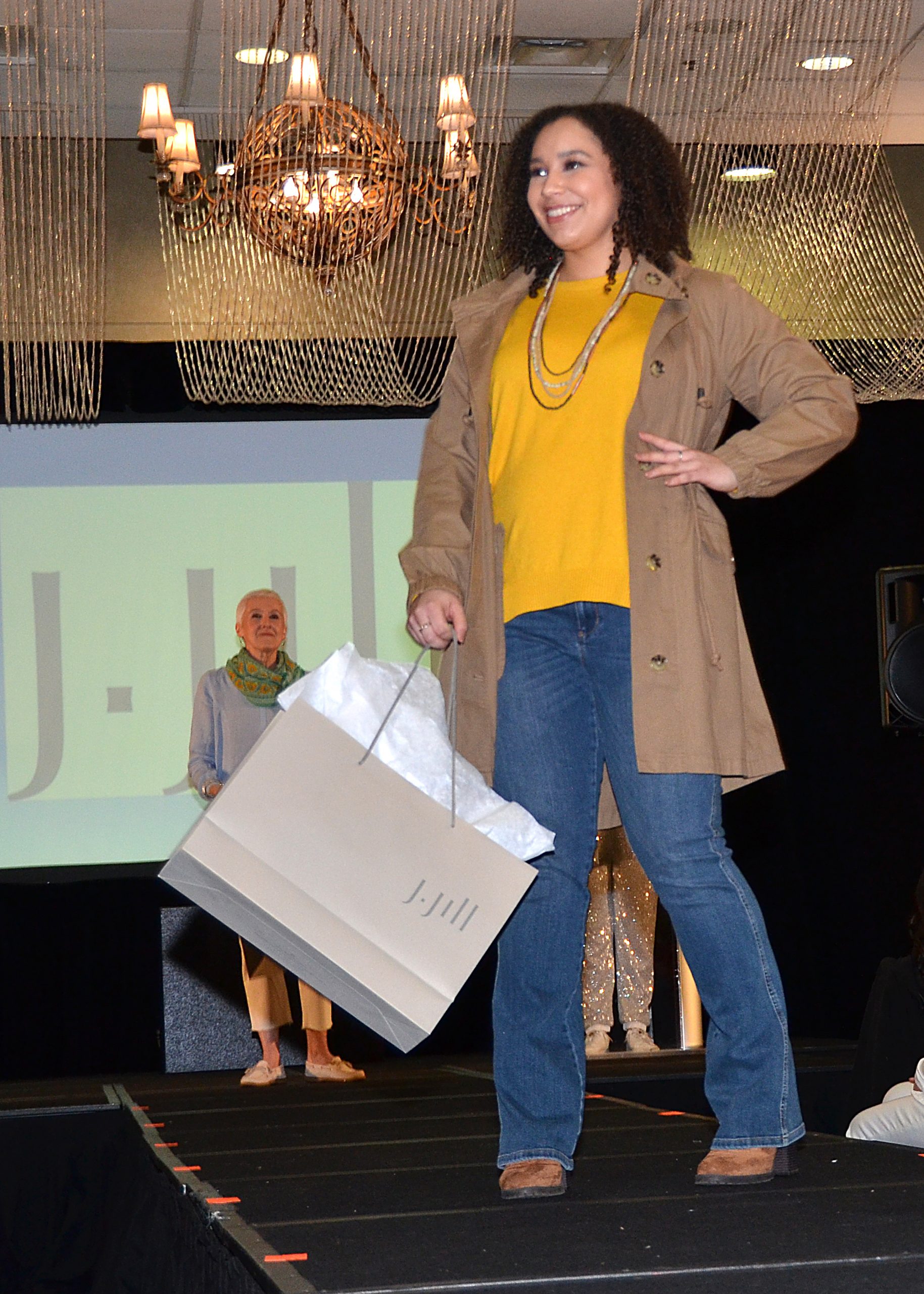 Model smiling on runway, holding shopping bag, wearing casual outfit.