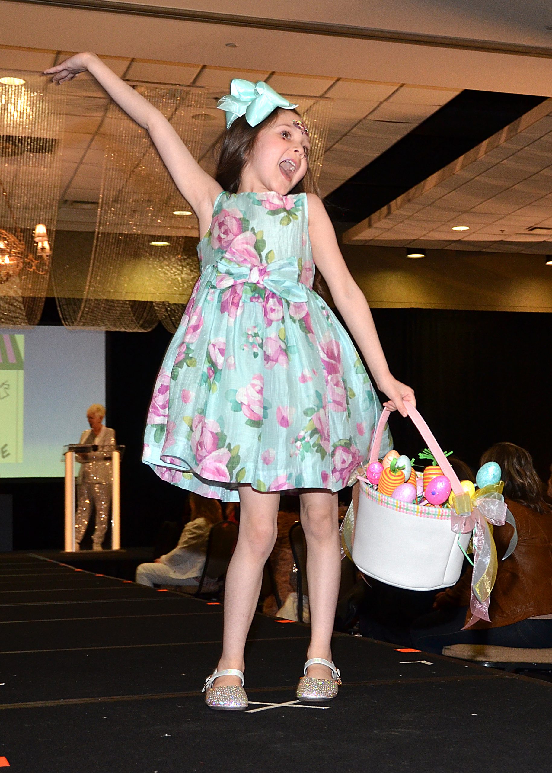 Child smiling on runway, holding basket of colorful eggs, wearing floral dress and bow.