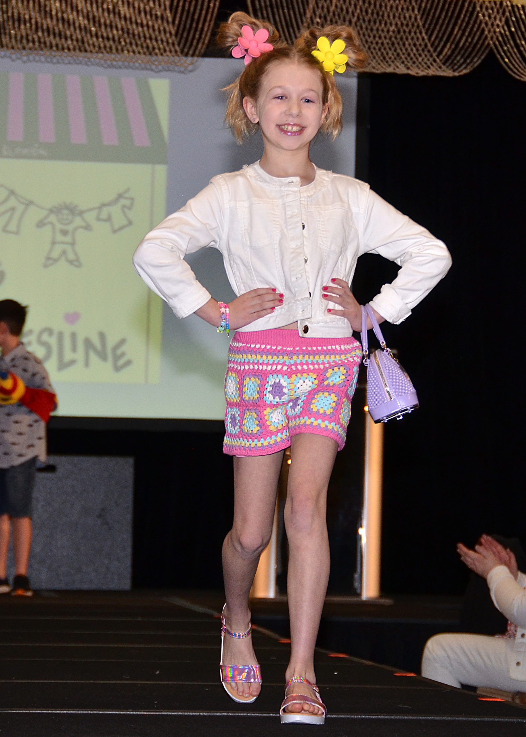 Young girl on runway, smiling, in colorful skirt and white top, with flowers in hair.