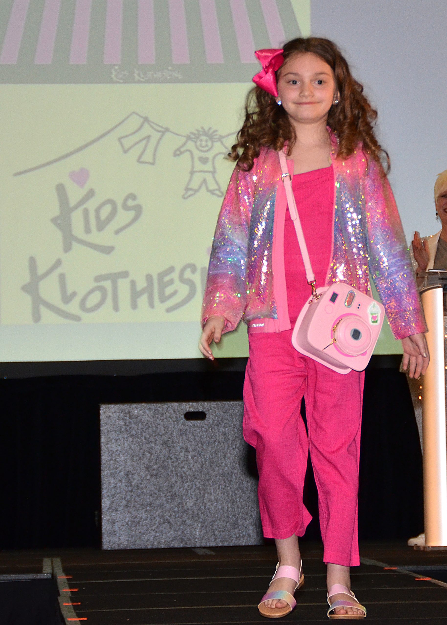 Young girl in pink and sparkly outfit walks on runway, smiling.