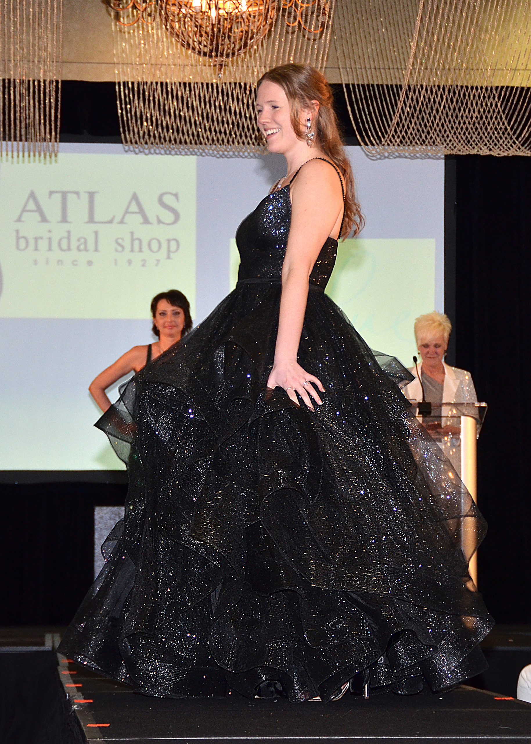 Woman in a black gown on a runway, smiling.