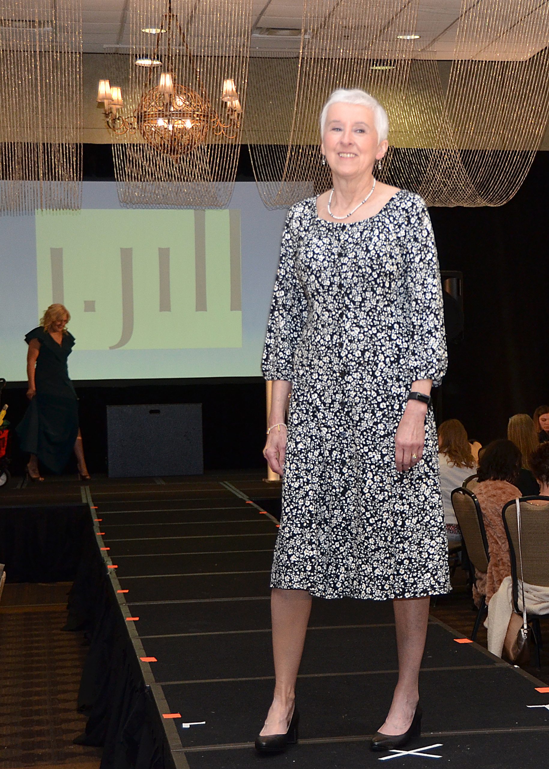 Senior woman in patterned dress on a runway, with audience seated nearby.