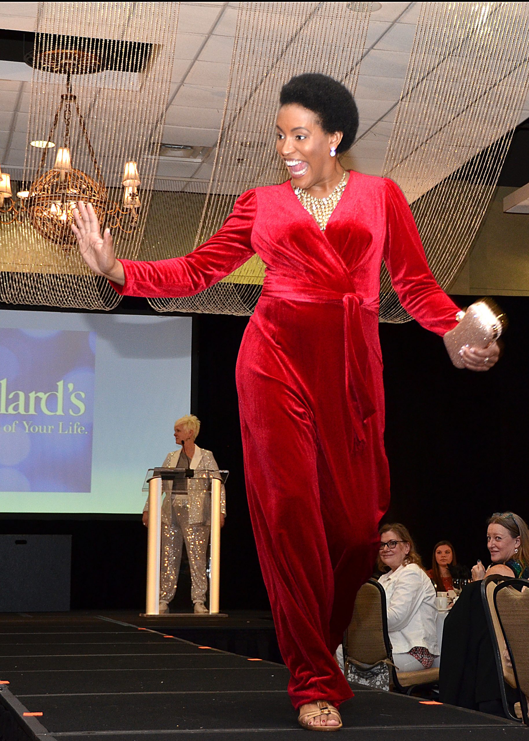 Model in red jumpsuit walks on runway, audience watches.