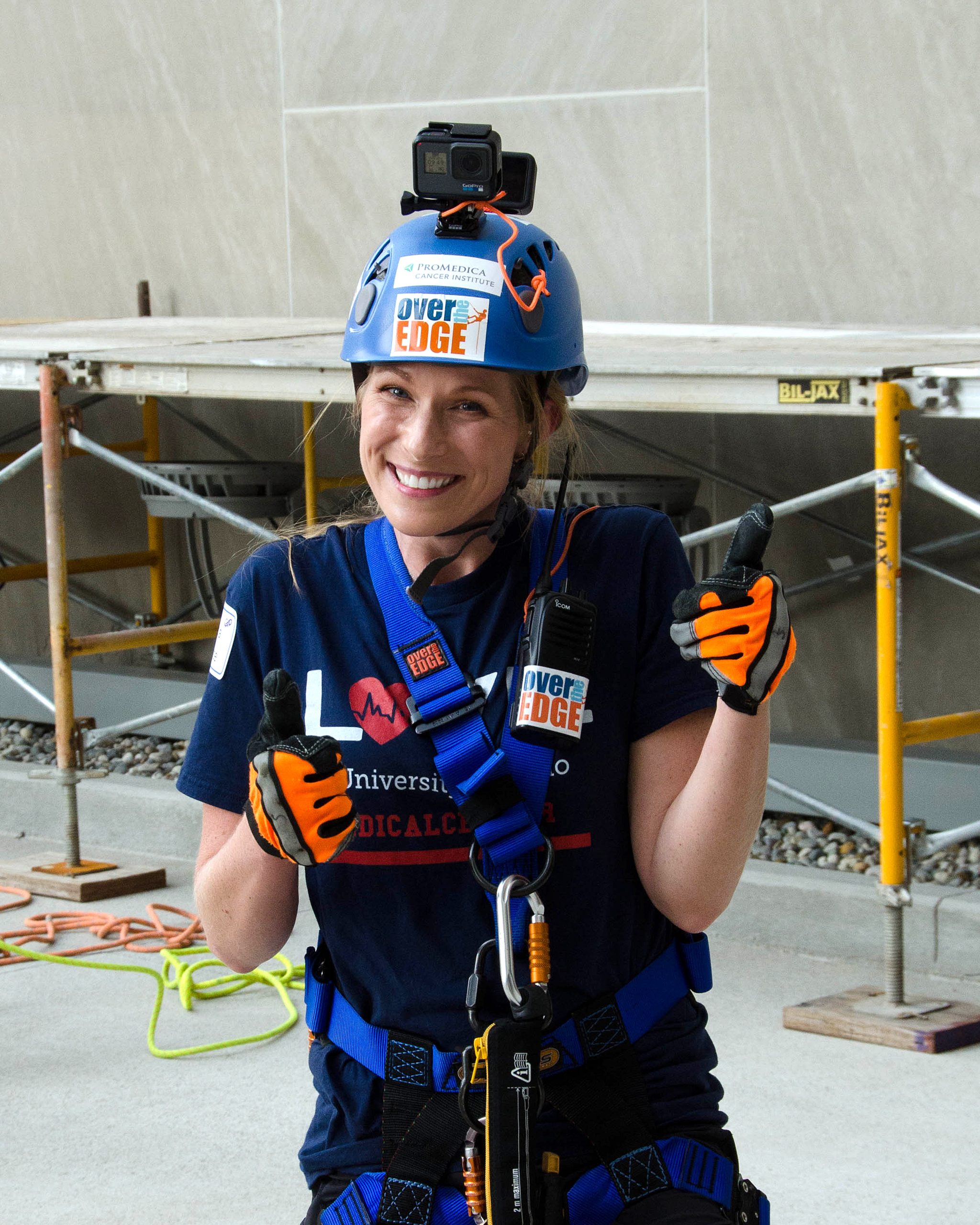 A person in climbing gear smiling and giving thumbs up indoors.