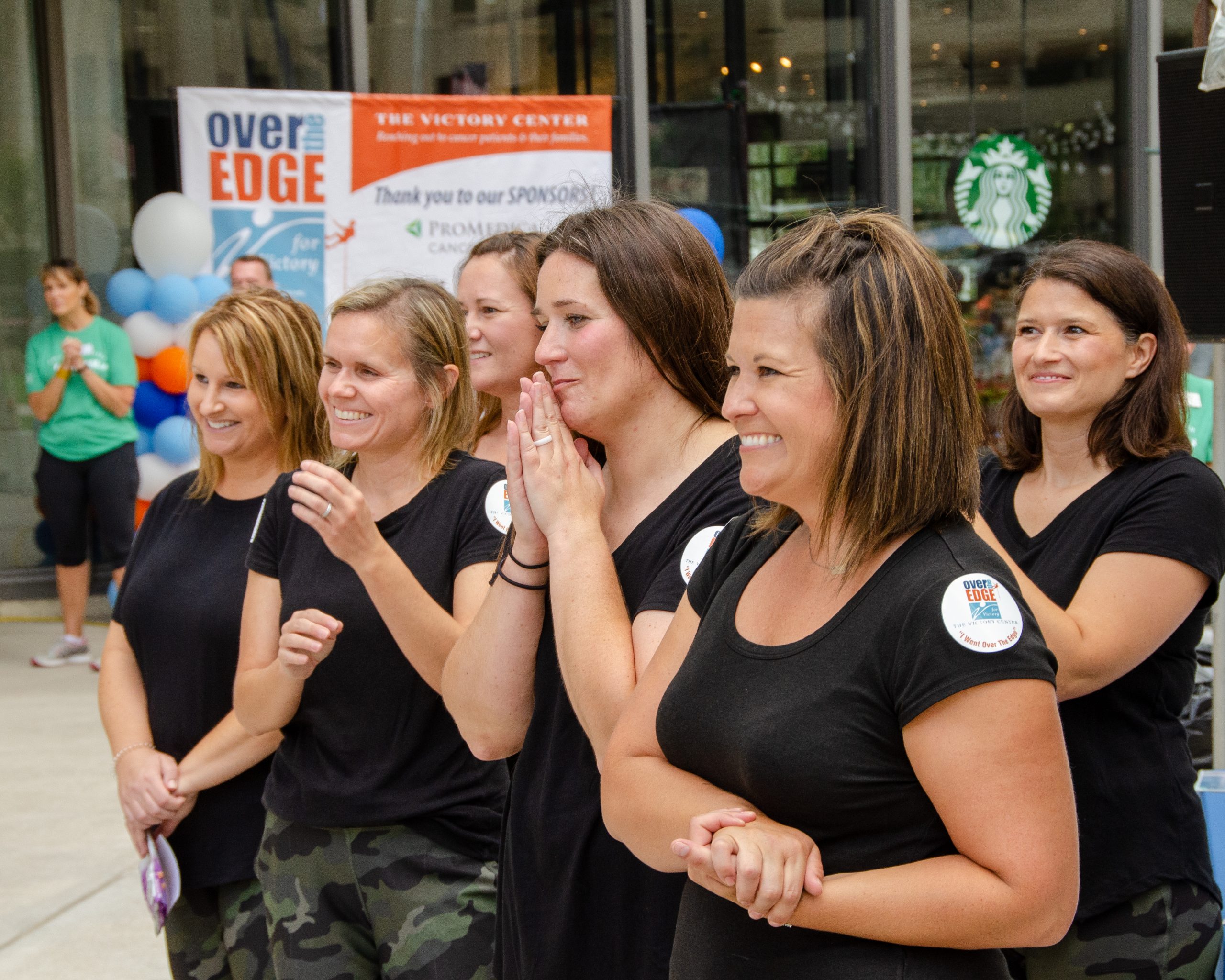 Women in black shirts clapping and smiling outdoors.