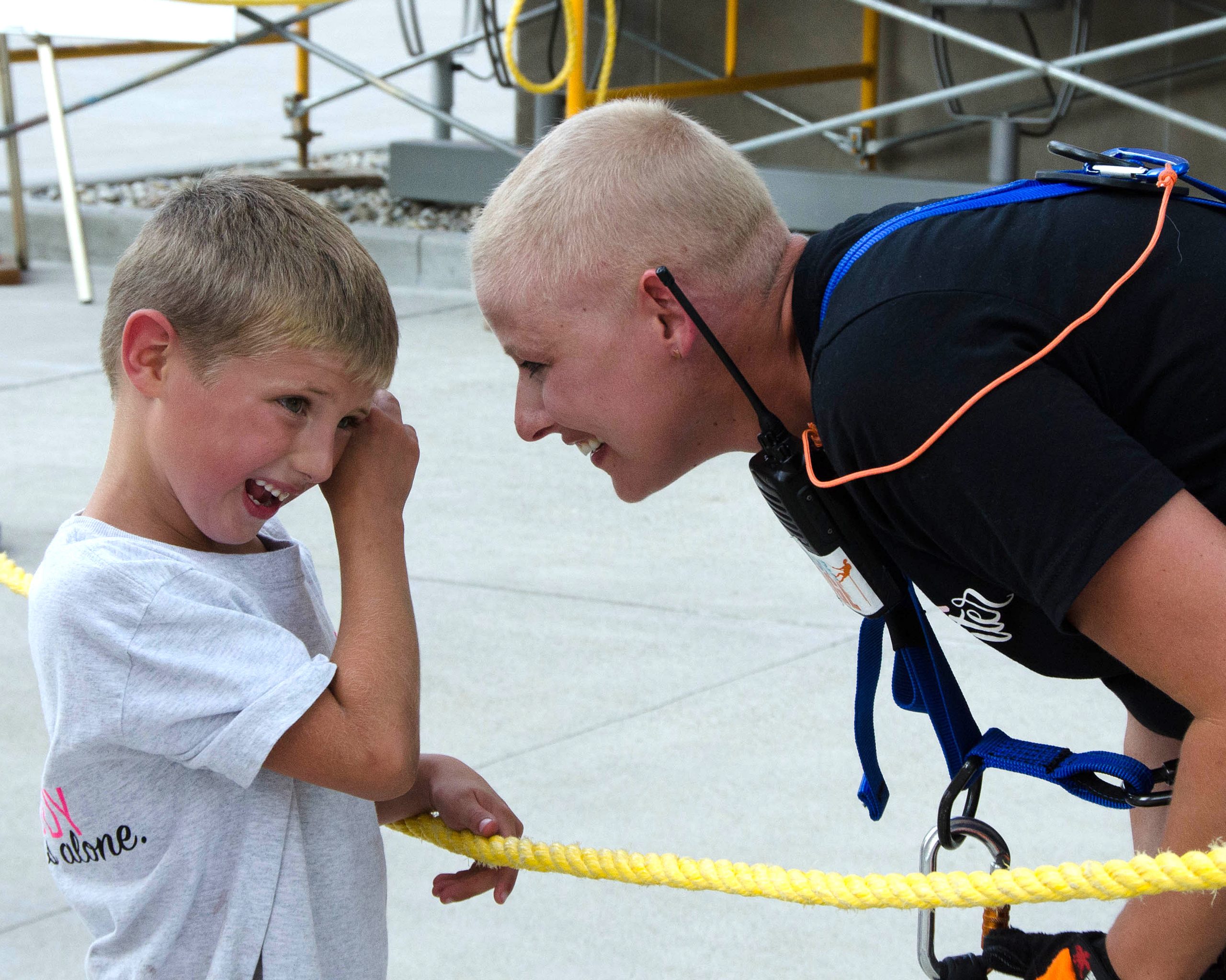 A smiling person in a harness leans toward a laughing child in a white shirt, separated by a rope.