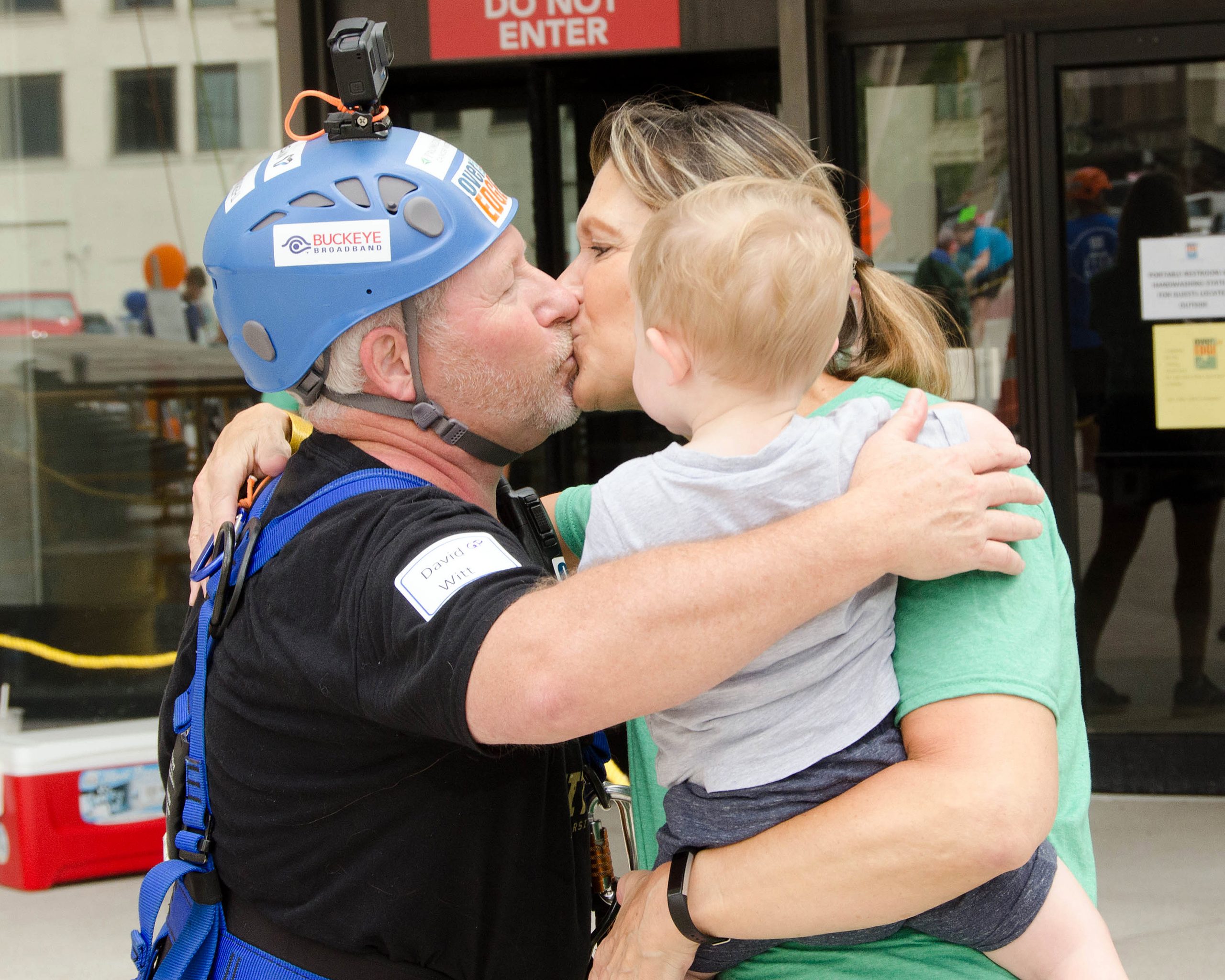 Man in climbing gear hugs and kisses woman holding a child.