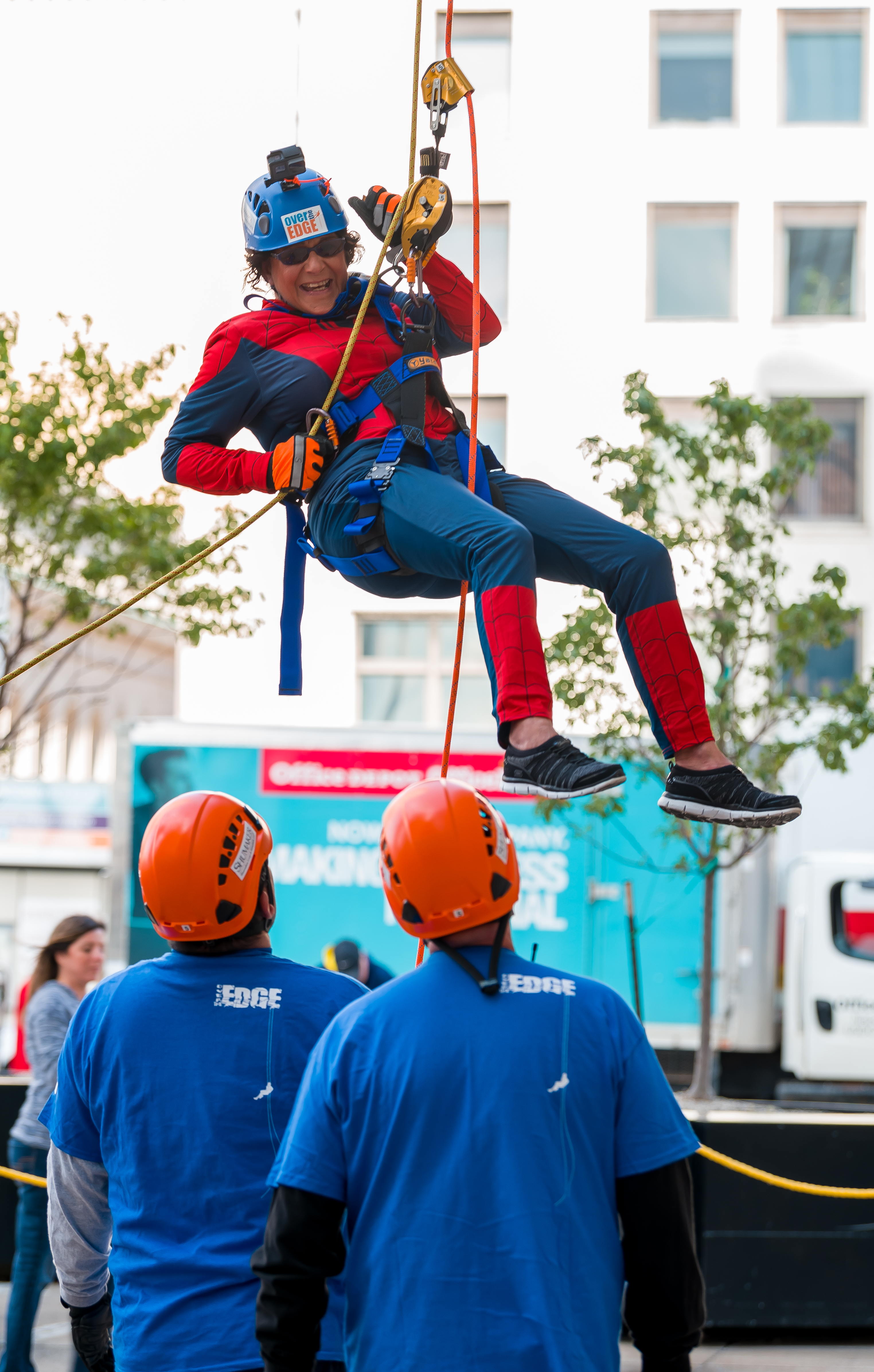 Person in superhero costume rappelling, watched by two people in blue shirts and orange helmets.