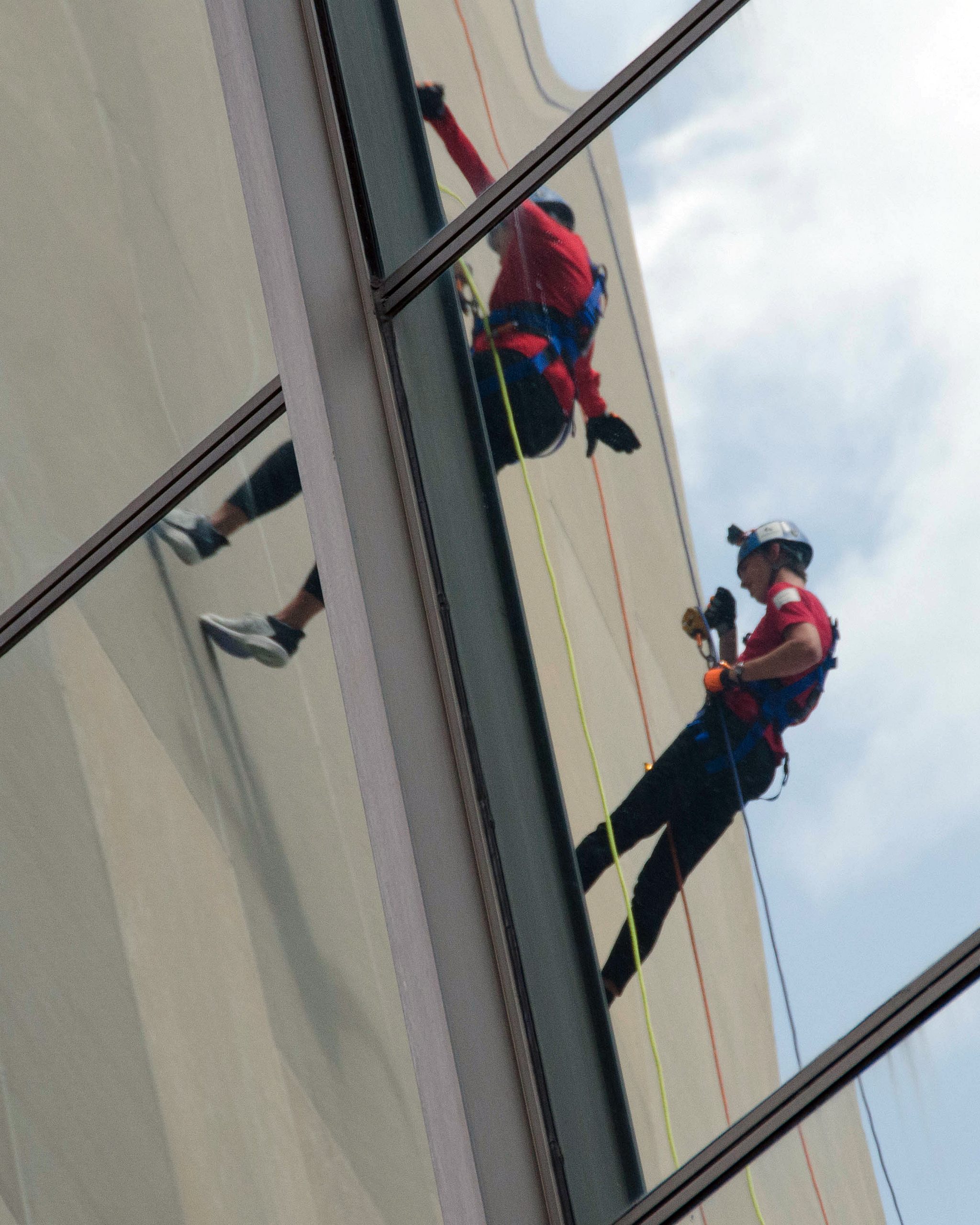 Window cleaner in red and black gear, reflected in glass building facade.