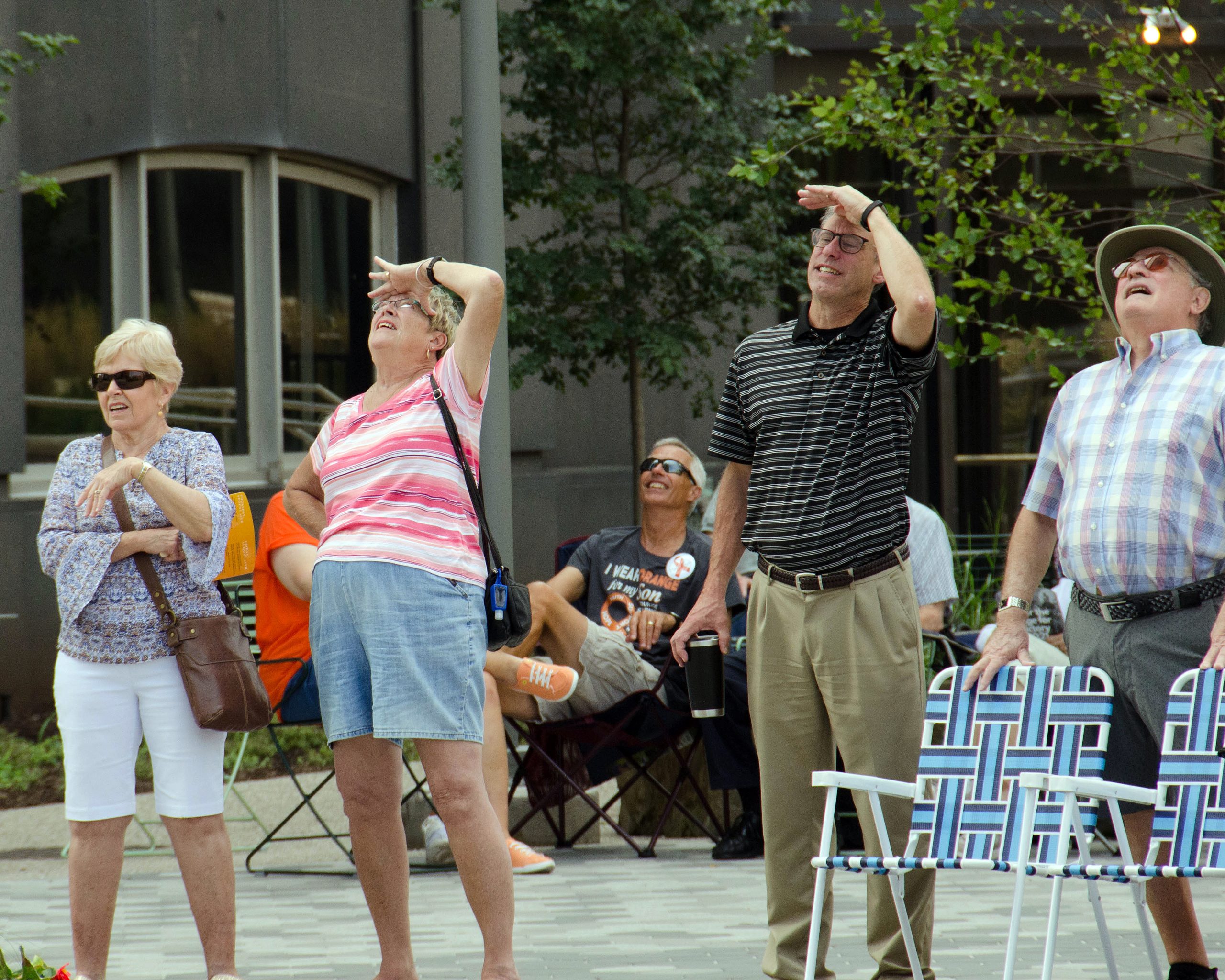 Four people looking up, shielding eyes from sunlight, with trees and buildings in the background.