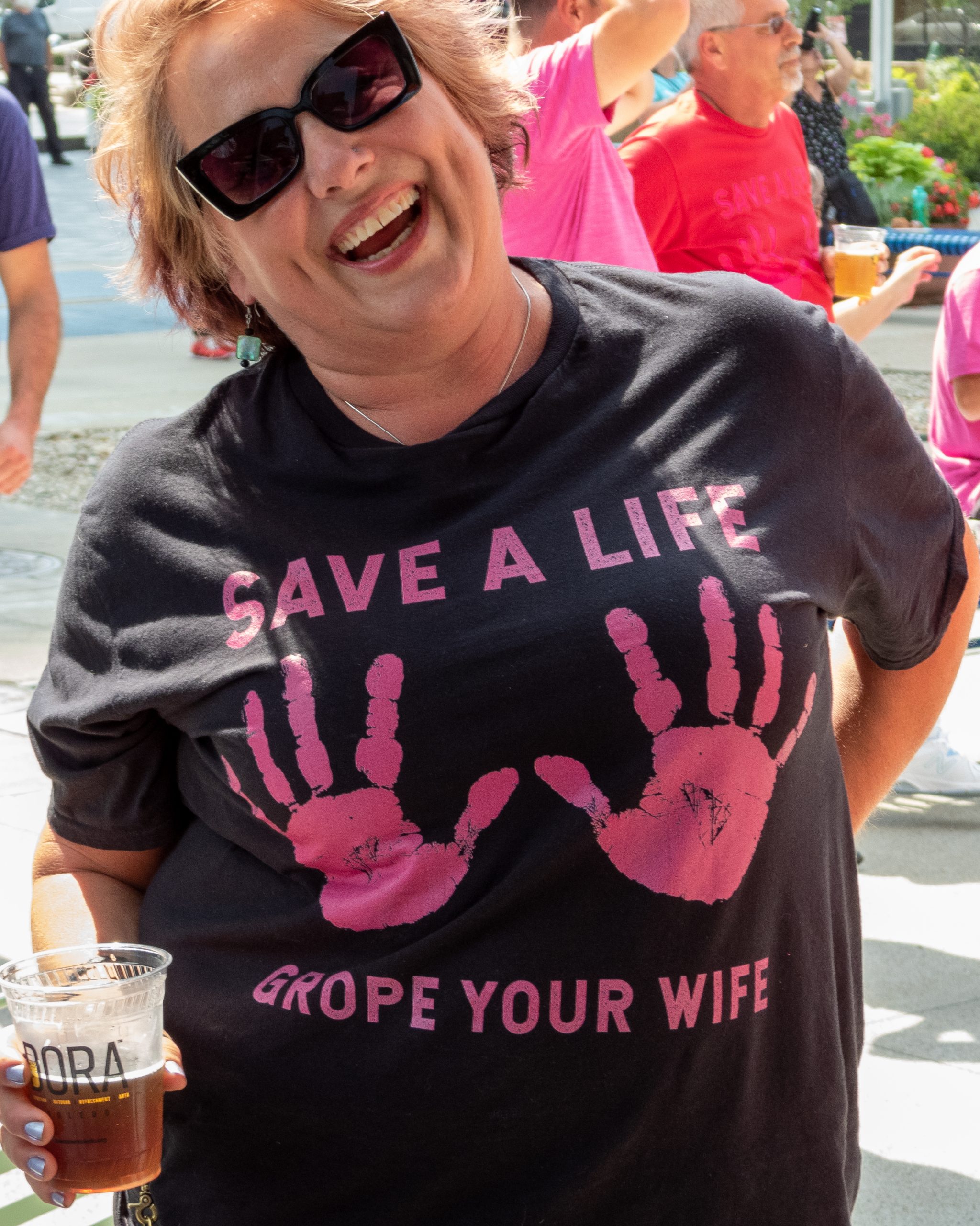 Woman wearing sunglasses and a humorous T-shirt, smiling outdoors with others nearby.