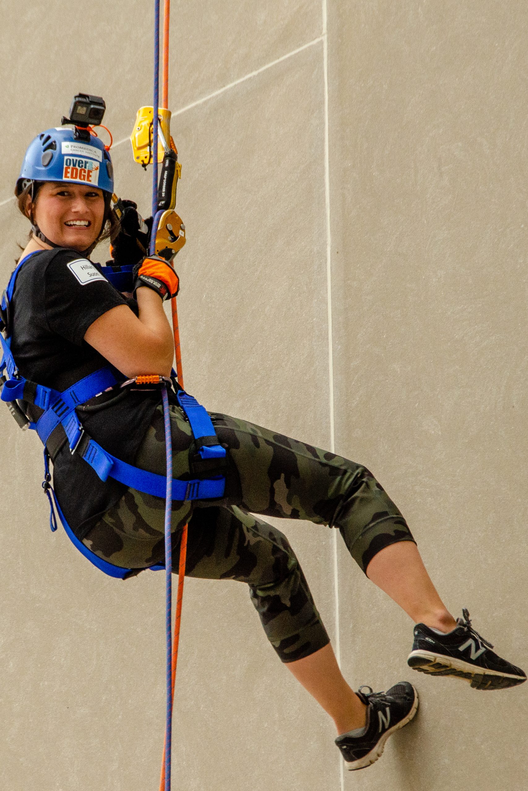 Person rappelling down a wall, wearing a helmet and harness, smiling.