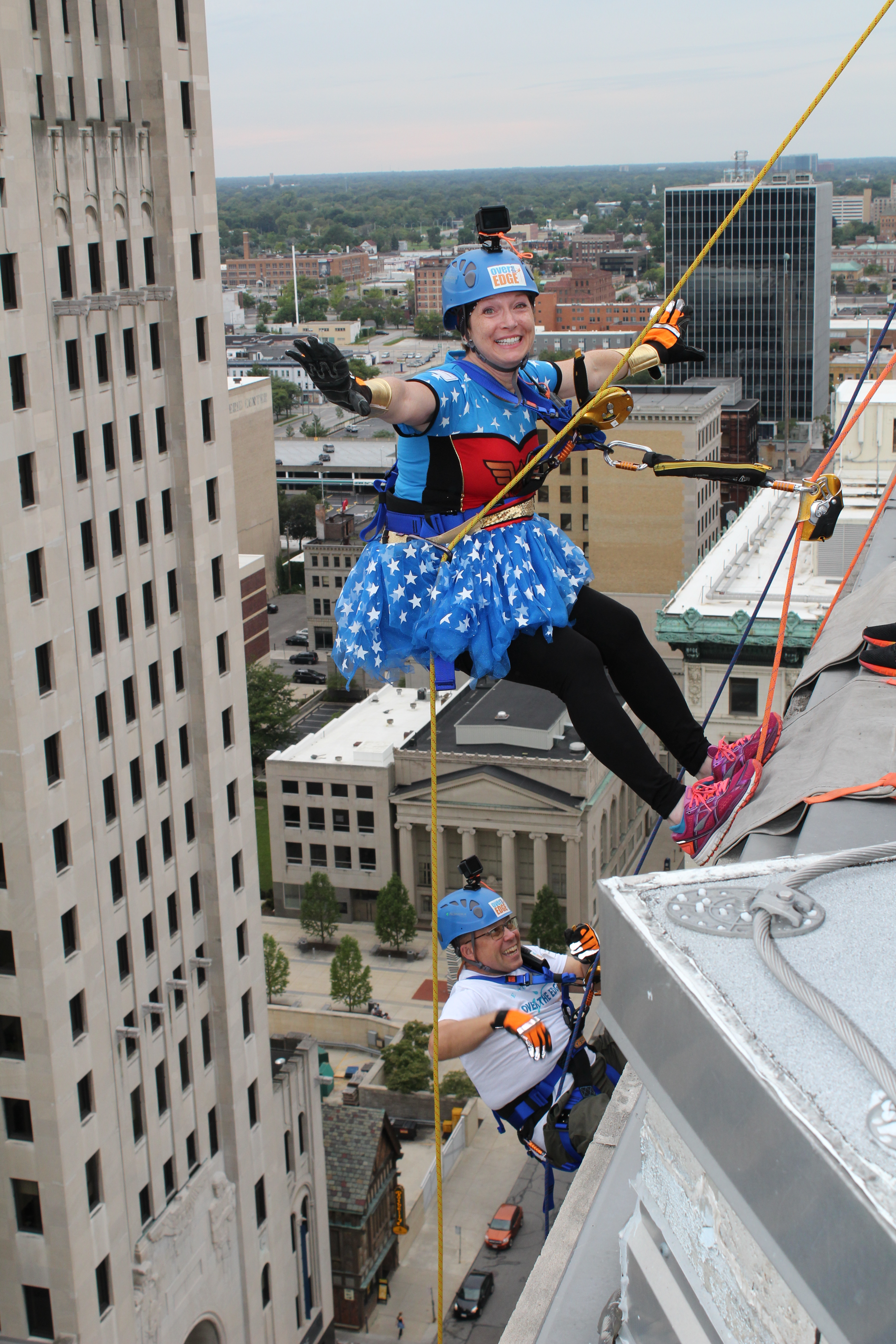Two climbers in costumes rappel down a tall building with a city view behind them.