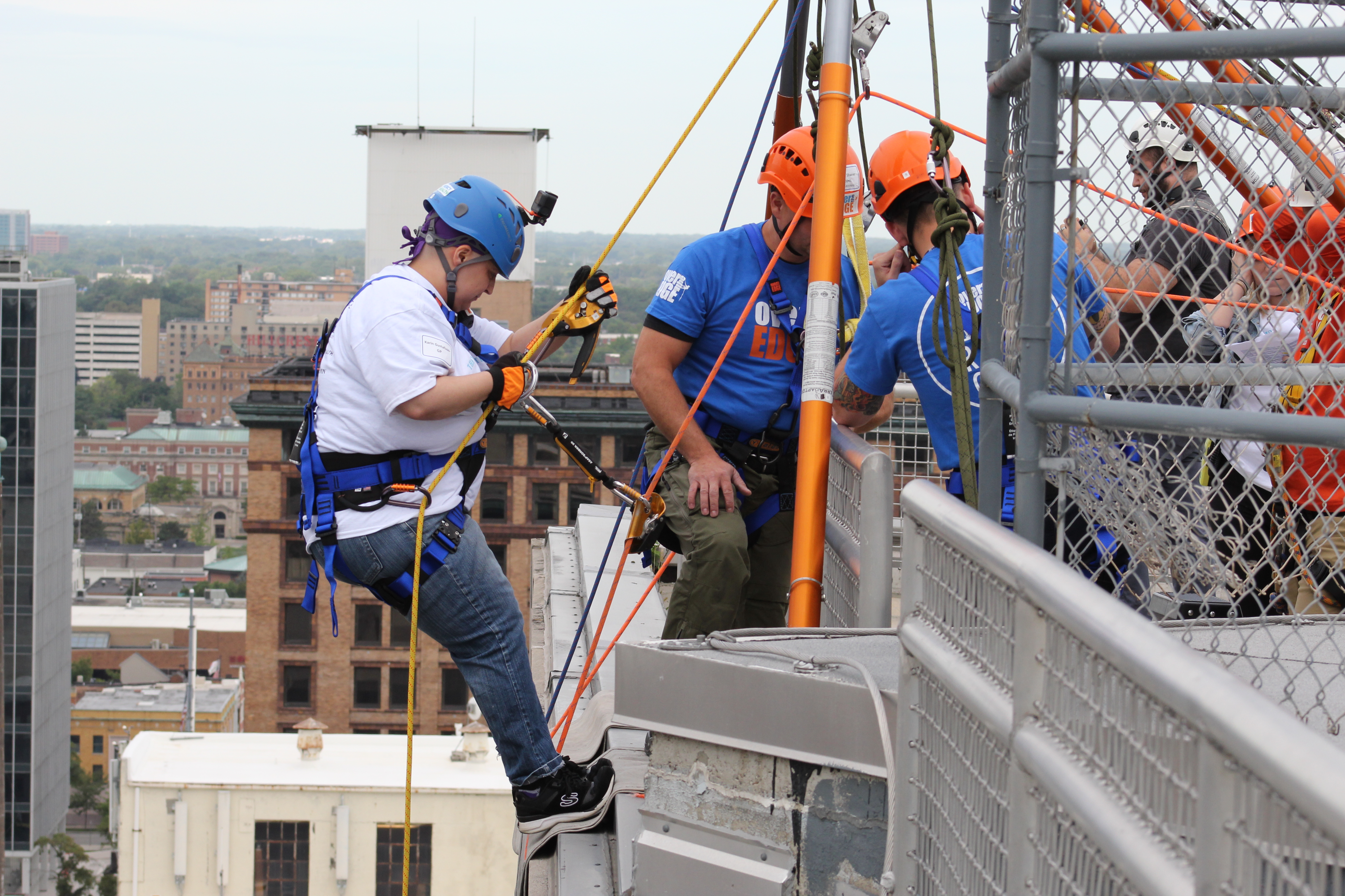 Person in harness rappelling down a building, assisted by workers in helmets. Urban background.