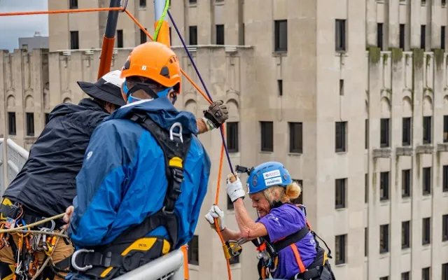 People in helmets and harnesses prepare to rappel down a tall building.