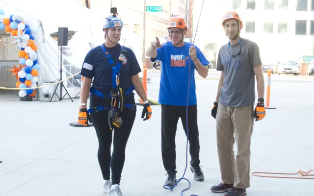 Three people in helmets and harnesses, standing on a city street, smiling.
