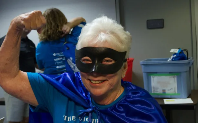 Person in blue cape and mask flexing arm confidently indoors.