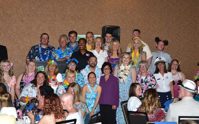 Group of people smiling, wearing colorful, tropical-themed attire in a banquet setting.