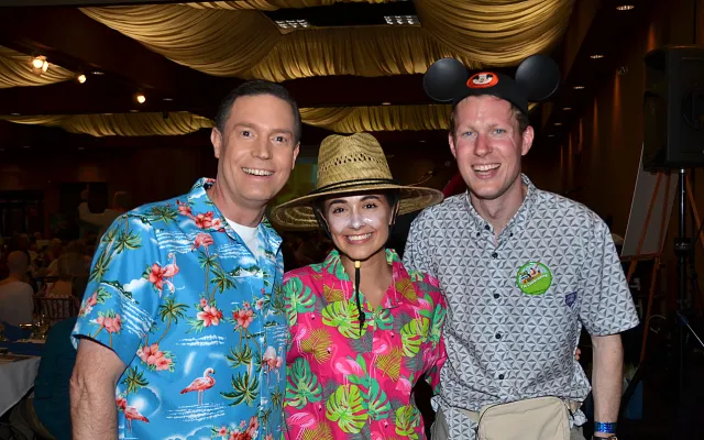 Three people in colorful shirts smiling; one wears a straw hat, another has mouse ears.