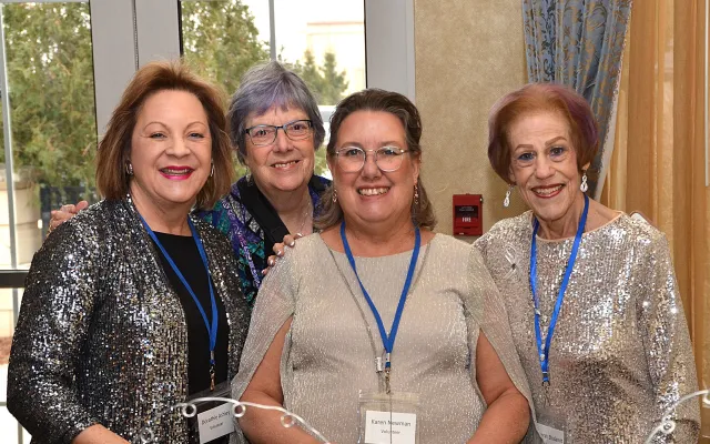 Four women smiling at an indoor event, wearing name tags and sparkly outfits.