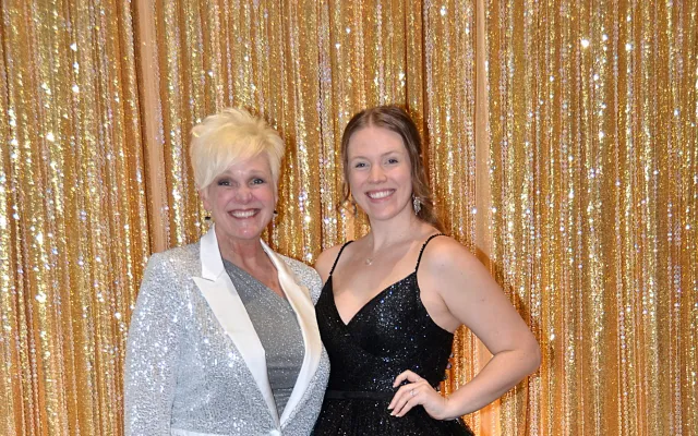 Two women smiling, one in a silver suit, the other in a black gown, standing before a gold curtain.