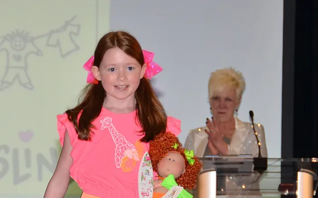 Young girl on stage with a doll. Woman clapping in the background.