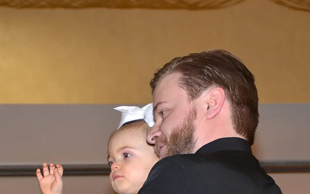 Man holding a baby with a bow, both gazing forward, indoors.