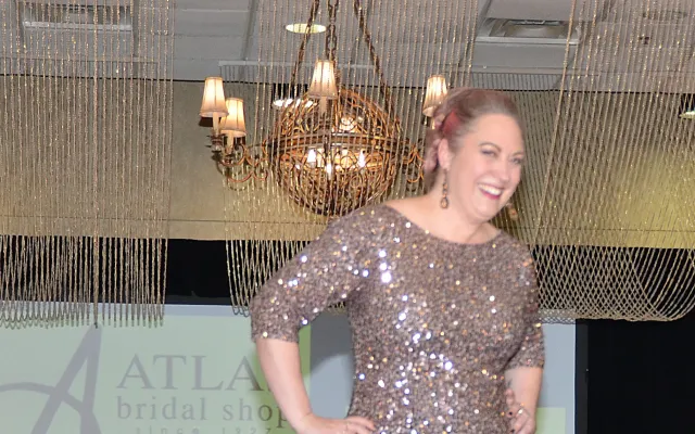 Model in a sparkling gown walks runway, others watch; chandelier overhead.