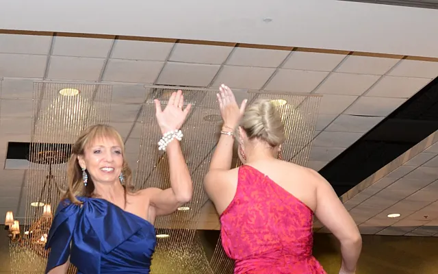 Two women high-fiving on a runway, wearing elegant blue and pink dresses.