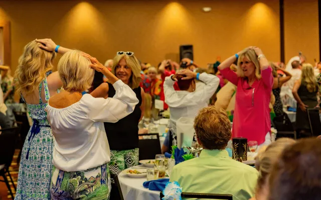 Women in a banquet hall dancing with hands on heads, colorful attire.