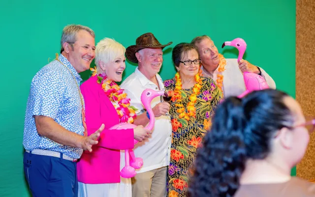 Five people in colorful attire pose with pink flamingos against a green background.