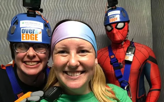 Three people smiling, one in a Spider-Man costume, wearing helmets and harnesses.