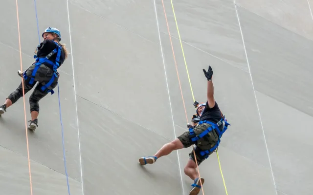 Two people rappel down a gray towering wall with colorful ropes.