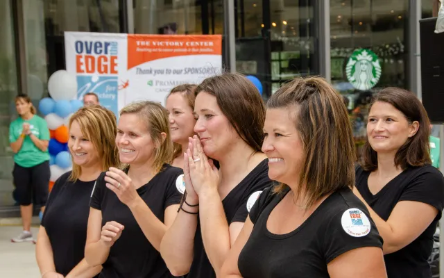 Women in black shirts clapping and smiling outdoors.