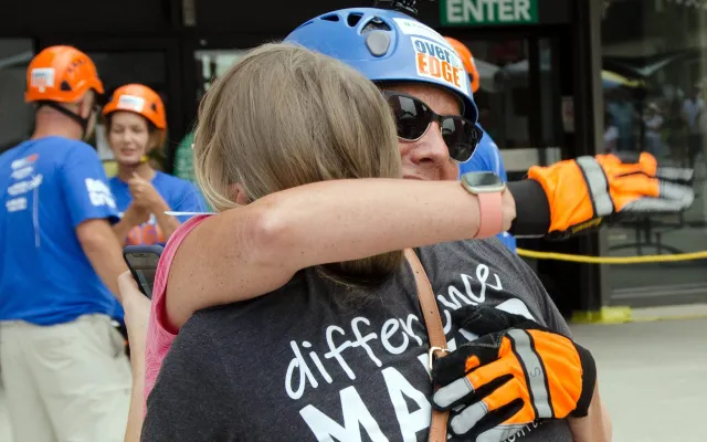 Two people wearing helmets hug outside, others with helmets in the background.