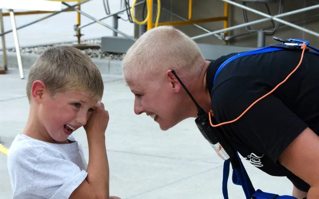 A smiling person in a harness leans toward a laughing child in a white shirt, separated by a rope.