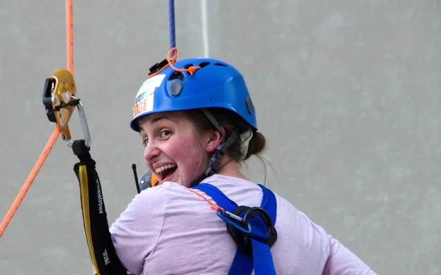 Woman with a blue helmet climbing, smiling at the camera.