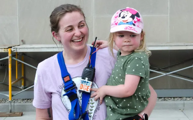 Woman in climbing gear holds a child wearing a Minnie Mouse hat, both smiling outdoors.