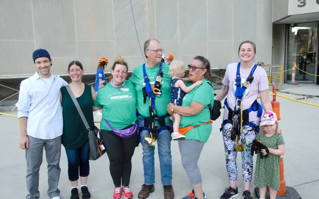 A group of smiling adults and children, some in climbing gear, standing together outdoors.