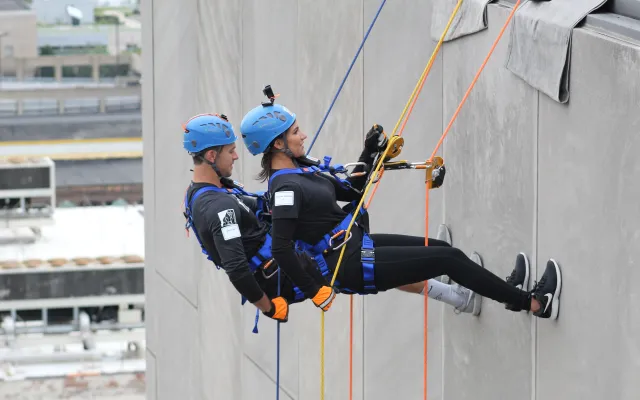 Two people in helmets and harnesses rappel down a building.
