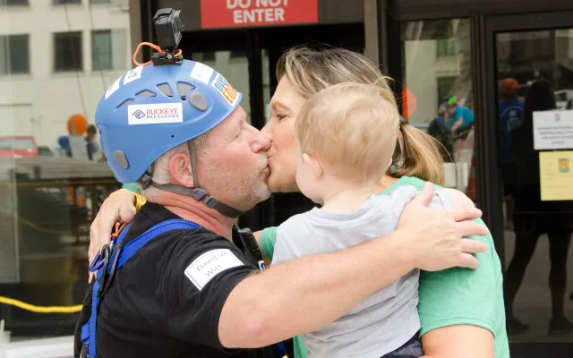 Man in climbing gear hugs and kisses woman holding a child.