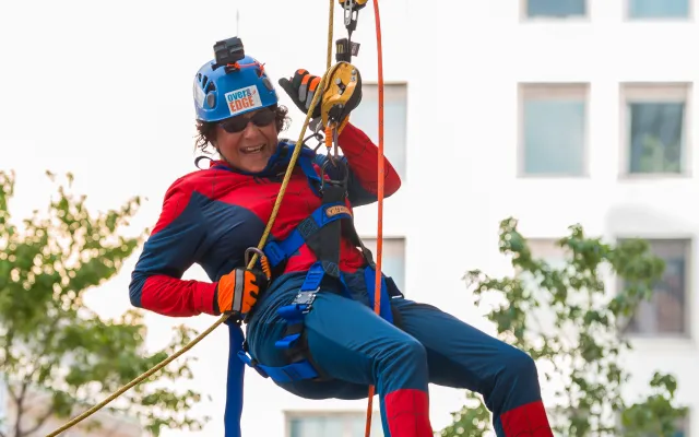 Person in superhero costume rappelling, watched by two people in blue shirts and orange helmets.