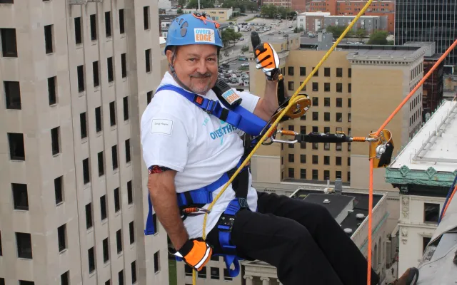 Person in safety gear rappels down a tall building, cityscape in the background.